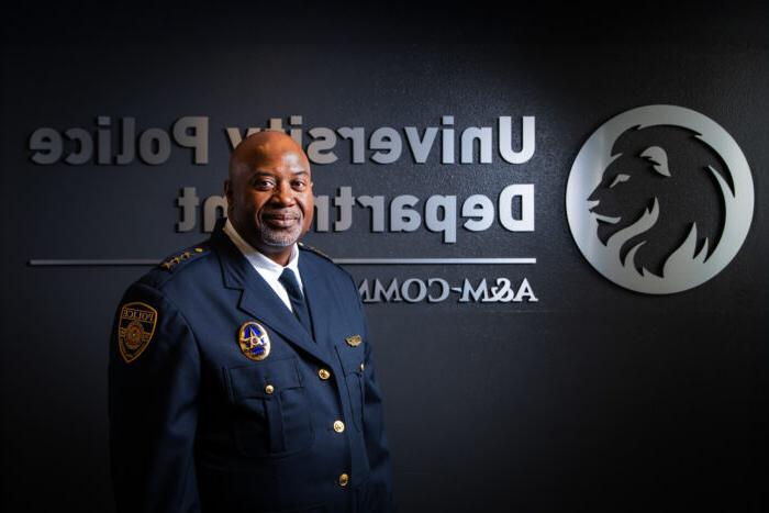 Chief Bryan Vaughn dressed in his dark blue, formal police uniform. He is standing in front of a dark wall with a silver embossed TAMUC lion head logo and the words 大学警察局 A&移动商务.