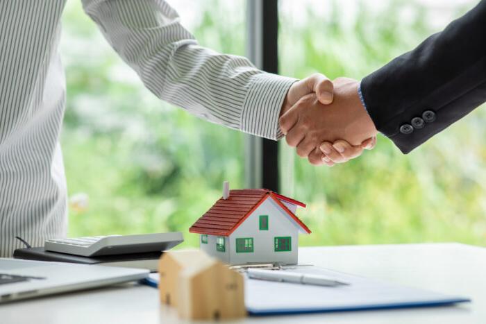 两个人握手, presumably over a real estate deal, with a table that contains a small model of a home beneath their hands.